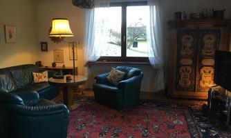 A cozy living room with blue leather sofas and a round table. The room is decorated with a carpet and a wooden cabinet, and a large window lets in plenty of light.