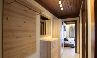 A bright hallway with wooden paneling, showing a wardrobe and a dresser. On the right side, a cozy living room is visible.