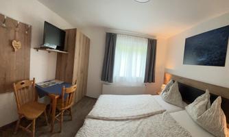 A bright room with a double bed, two chairs, and a table. A television hangs on the wall and a window provides daylight.