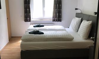 A modern bedroom with two twin beds and white bedcovers. There are large windows with curtains and a light wooden floor.