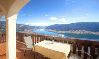 A beautiful view from a terrace over a lake and surrounding mountains. On the table is a bottle of wine and there are two chairs.