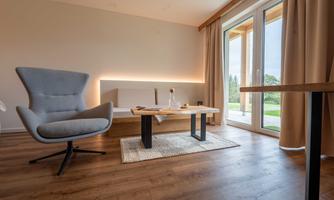 A modern room with a comfortable armchair and a wooden table. Large windows let in plenty of daylight and offer views of nature.