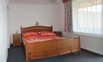 A simple bedroom with a wooden bed and red bedcovers. Large windows provide plenty of daylight.