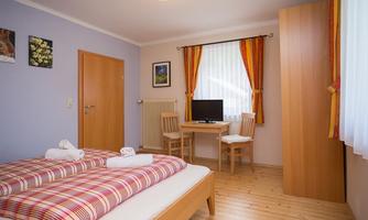 A cozy bedroom with two beds and wooden floors. The room is bright and features a table, chairs, and windows with curtains.