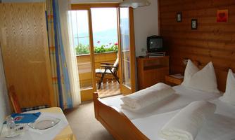 A cozy room with wooden-paneled walls and a double bed. In the background, there is a balcony overlooking nature.