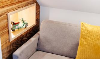 A cozy sofa with a yellow cushion. On the wall, there is a picture of a green Vespa in a niche.