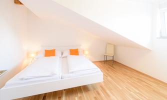 A bedroom with a sloped roof and a bed in the center. There are bright walls, a wooden floor, and orange cushions on the bed.