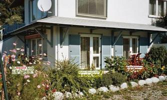 A bright house with blue shutters and an inviting terrace. In front of the house, a colorful garden with many flowers is growing.