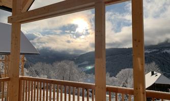 View of a snow-covered landscape from a wooden porch. In the background, mountains and a cloudy sky with the sun can be seen.