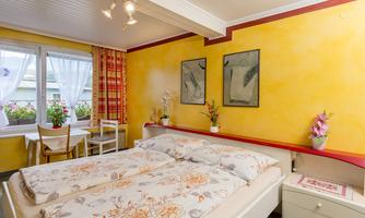A cozy bedroom with a large bed and bright walls. The decor is simple and welcoming, with a table next to the window.