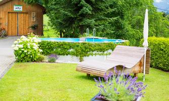 A well-maintained garden with a lounger and a blooming lavender bush. In the background, there is a swimming pool and a wooden house surrounded by green trees.