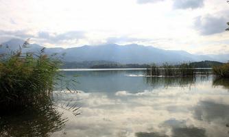 A calm lake, surrounded by mountains and reeds. The sky is cloudy and reflected in the water.