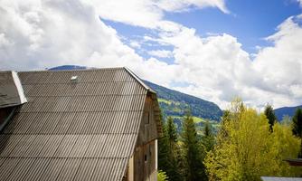 A rural scene with a wooden building and a green mountain in the background. The sky is partly cloudy and features beautiful cloud formations.