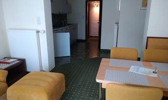 A cozy living room with a table and yellow armchairs. In the background, a kitchen and a hallway can be seen.
