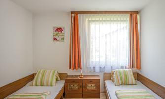 A simple bedroom with two single beds and colorful bedspreads. Bright curtains hang in front of the window, and there is a small nightstand.