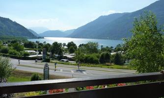 A picturesque landscape with a quiet lake and high mountains in the background. The surroundings are green and offer a view of motorhomes and a road.