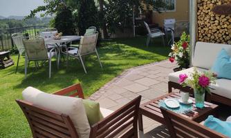 An inviting garden lounge with cozy wooden furniture and colorful flowers. In the background, there are tables and chairs for social gatherings.