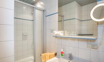 A modern bathroom with a shower, sink, and a large mirror. The walls are light tiled, and there are towels and toiletries.