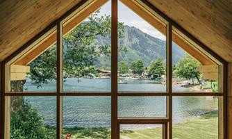 A view from a wooden house over a peaceful lake, surrounded by trees and mountains. The sky is clear and the landscape looks inviting.