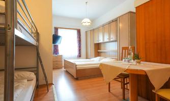 A bright, cozy room with two beds, including a bunk bed. A table with chairs and large windows create an inviting atmosphere.