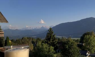 A glass of beer on a wooden table with a view of mountains and the clear sky. In the background, trees and a tranquil landscape can be seen.