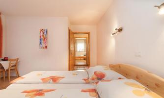 A cozy bedroom with two beds and colorful bed covers. In the background, a table and an open room can be seen.