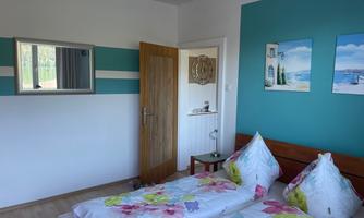 A bright bedroom with two beds and colorful bedding. The walls are painted in soft blue and white tones, with pictures by the window.