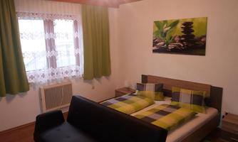 A cozy bedroom with a double bed and green decor. Large windows let in plenty of light, and a piece of artwork featuring stones and plants adorns the wall.