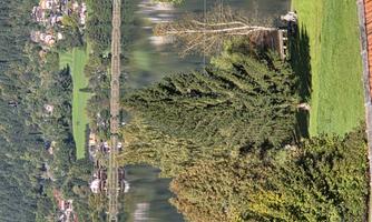 A peaceful lake surrounded by trees and a green meadow. In the background, gentle hills and houses can be seen.