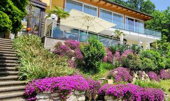 A charming house with a beautiful balcony and large windows. The garden is adorned with colorful flowers and green plants.
