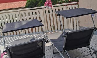A balcony with two loungers and a sun umbrella. In the background, green trees and a red roof are visible.