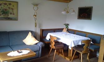 A cozy living room with a gray sofa, a wooden table, and chairs. Pictures hang on the walls, and a table is decorated with a tablecloth.