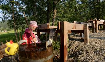 A child is playing in a wooden basin with water and has a red rubber duck. In the background, there are trees and a wooden structure visible.