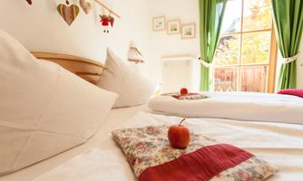 A cozy bedroom with two beds and a warm atmosphere. On the bedspread lies a red apple and there are green curtains that let in light.