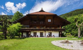 A traditional wooden house surrounded by green meadows and trees. The sky is blue with some white clouds.
