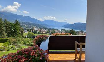 A balcony with colorful flowers and a view of the mountains and a lake. The sky is clear and blue, the landscape is green and inviting.