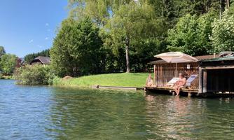 A calm lake with a green shore and trees. Two people are relaxing on a platform by the water.