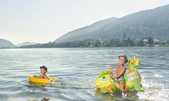 Two children are playing in the water. One is sitting in a yellow swimming ring, while the other is swimming on an inflatable turtle toy.