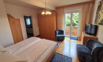 A cozy bedroom with a double bed, two armchairs, and a television. Large windows offer a view of the balcony and the green surroundings.