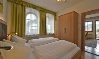 A cozy bedroom with two beds and large windows. The walls are light, and the curtains are green.