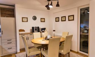 A modern dining room with a round wooden table and light chairs. Pictures are hanging on the walls, and there are windows with a view outside.