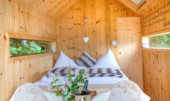 A cozy wooden cabin with a double bed and space for two chairs. The room is bright and inviting, with many windows and a small table with flowers.
