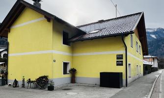 A yellow building with a gabled roof in a rural setting. The sky is gray and a quiet road is visible.