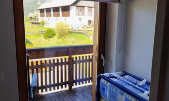 A view from a room onto a balcony with a wooden railing. In the background, green meadows and mountains can be seen.