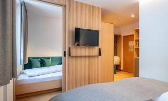 A modern hotel room with wooden paneling and a television on the wall. In the background, another room can be seen with a cozy bed.
