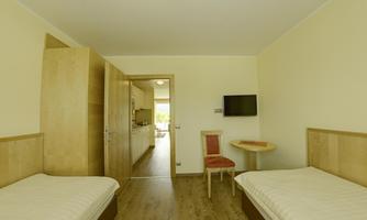 A bright room with two single beds, a table, and a television. The walls are painted in soft colors and a window leads outside.