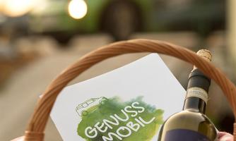 A basket with a wine bottle and a card that says "GENUSS MOBIL". In the background, a green car is visible.