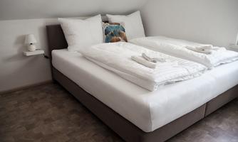 A modern bedroom with a large double bed and fresh towels. The walls are light, and a bedside table with a lamp is available.
