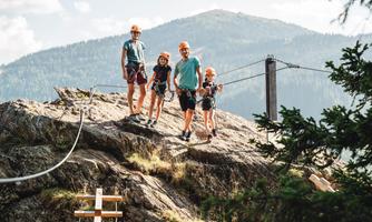 A family is standing on a rock in nature, ready to climb. In the background, mountains and a clear sky are visible.
