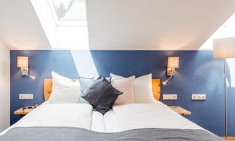 A cozy bedroom with two beds and bright pillows. The walls are blue, and there is a stylish lamp as well as daylight from a skylight.